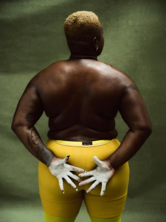Back view of plus-size adult female with light blonde buzz cut wearing yellow shorts, hands painted white, against muted green backdrop.