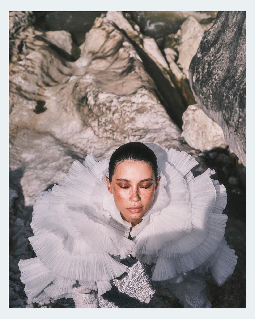 Woman with closed eyes wears a ruffled white collar surrounded by gray rocks. The shot features soft lighting and a serene atmosphere.