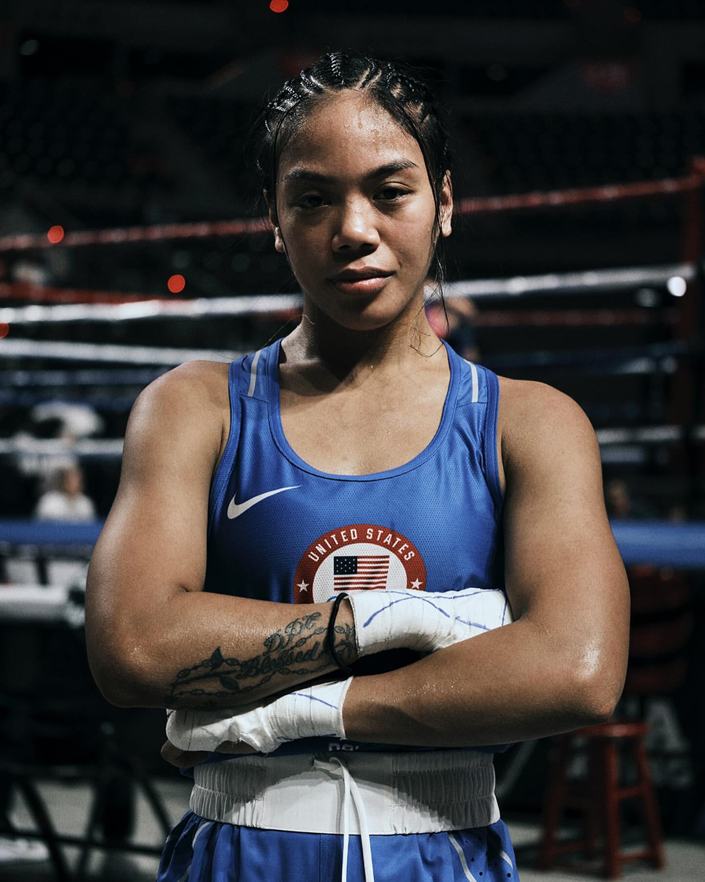 Female boxer with braids in a blue tank top with the United States flag, posing in the boxing ring with arms crossed, serious expression.