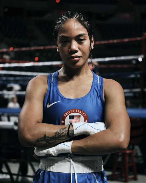 Female boxer with braids in a blue tank top with the United States flag, posing in the boxing ring with arms crossed, serious expression.