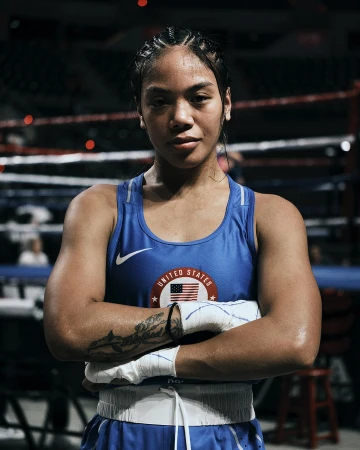 Female boxer with braids in a blue tank top with the United States flag, posing in the boxing ring with arms crossed, serious expression.