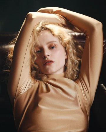 Portrait of a blonde woman in a gold high-neck top with arms raised, photographed by Sharif Hamza.