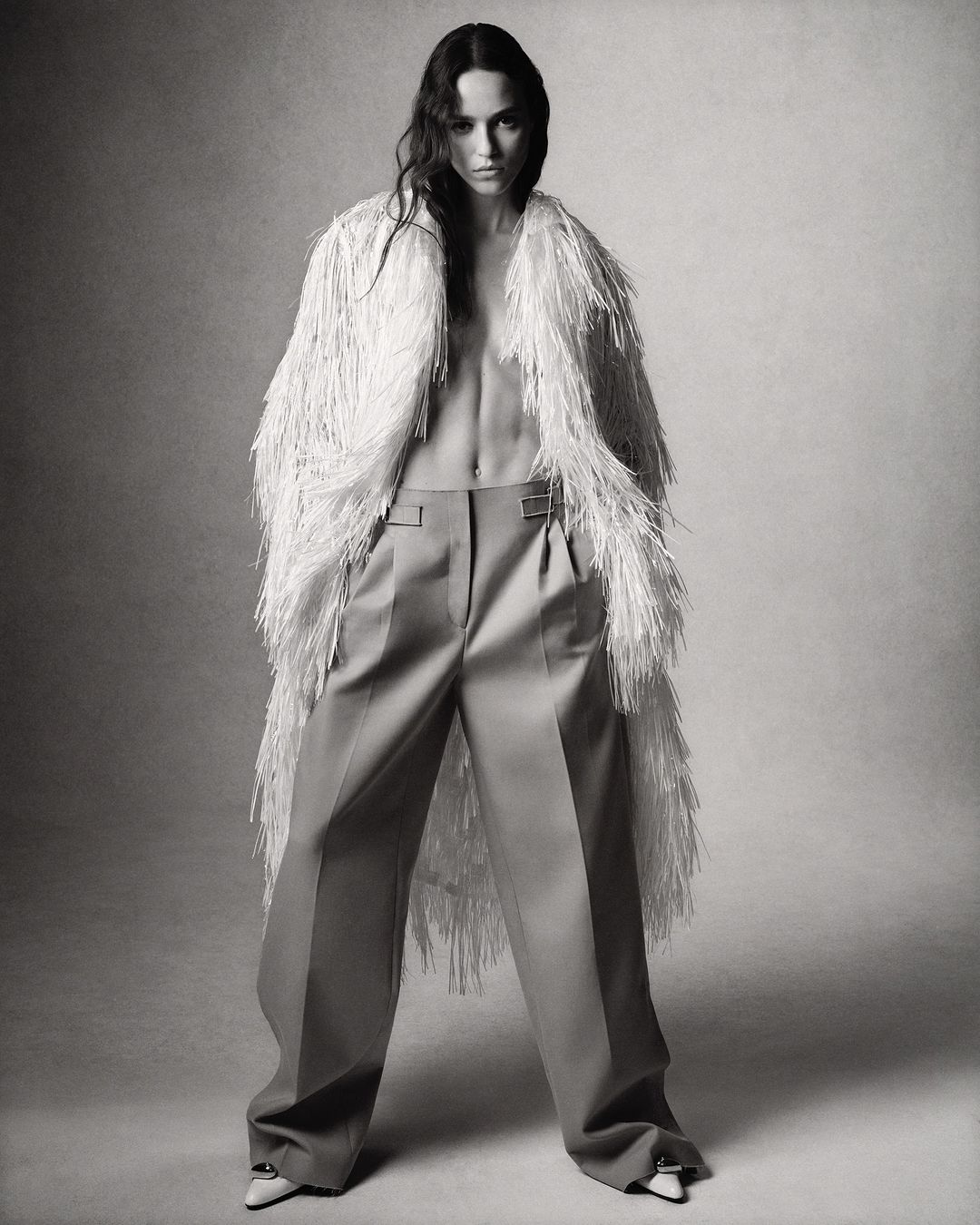 Fashion shot of a woman in wide-leg pants and a fringed coat, posing in a studio with medium shot and monochromatic palette.