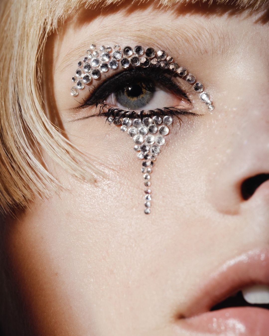 Close-up portrait of a fair-skinned adult female with blonde hair and intricate rhinestone eye makeup. Studio lighting and shallow depth of field.