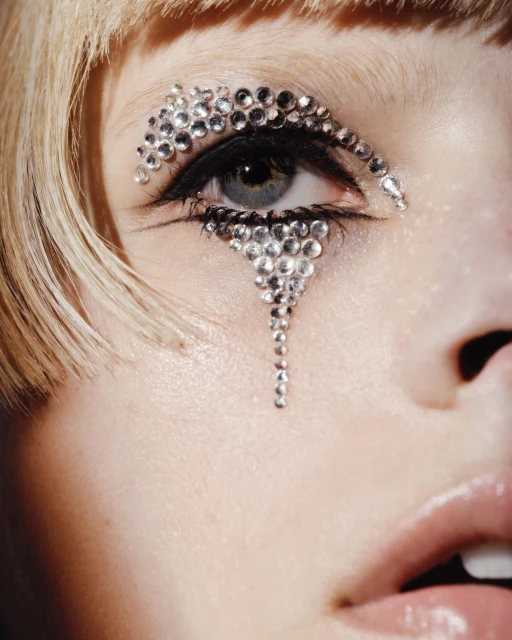Close-up portrait of a fair-skinned adult female with blonde hair and intricate rhinestone eye makeup. Studio lighting and shallow depth of field.