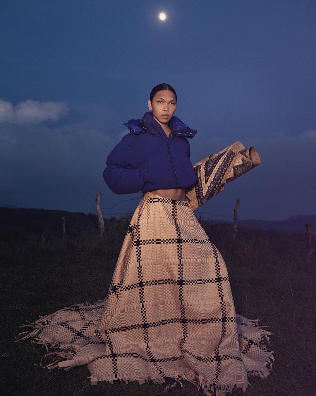 A person standing in a field at night, wearing a puffy jacket and a woven skirt. The moon is visible in the night sky.