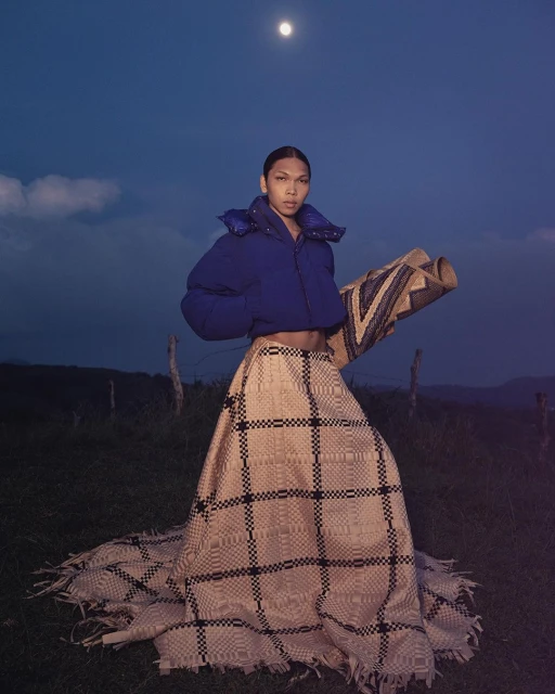 A person standing in a field at night, wearing a puffy jacket and a woven skirt. The moon is visible in the night sky.
