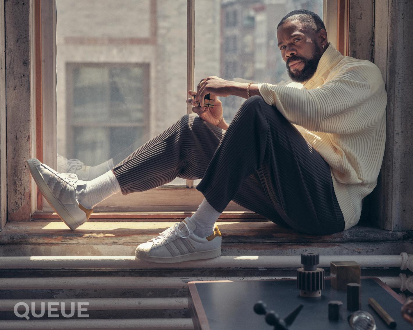 Adult male poses in a window wearing a light yellow top, brown pleated pants and Adidas sneakers, with geometric props and diffused light.