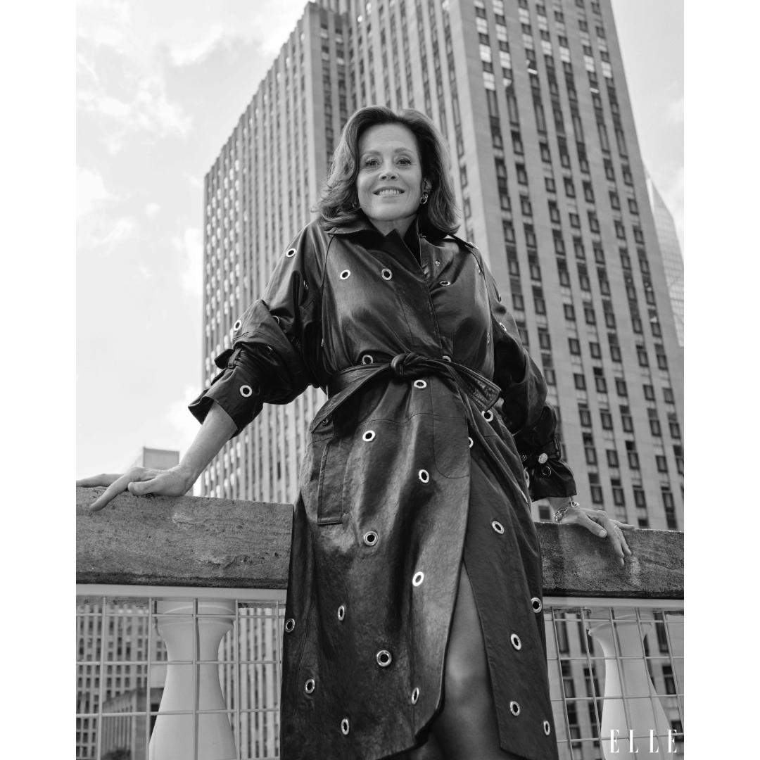 Black and white portrait of Sigourney Weaver in a black leather coat in front of a city building with cloudy sky in the background.