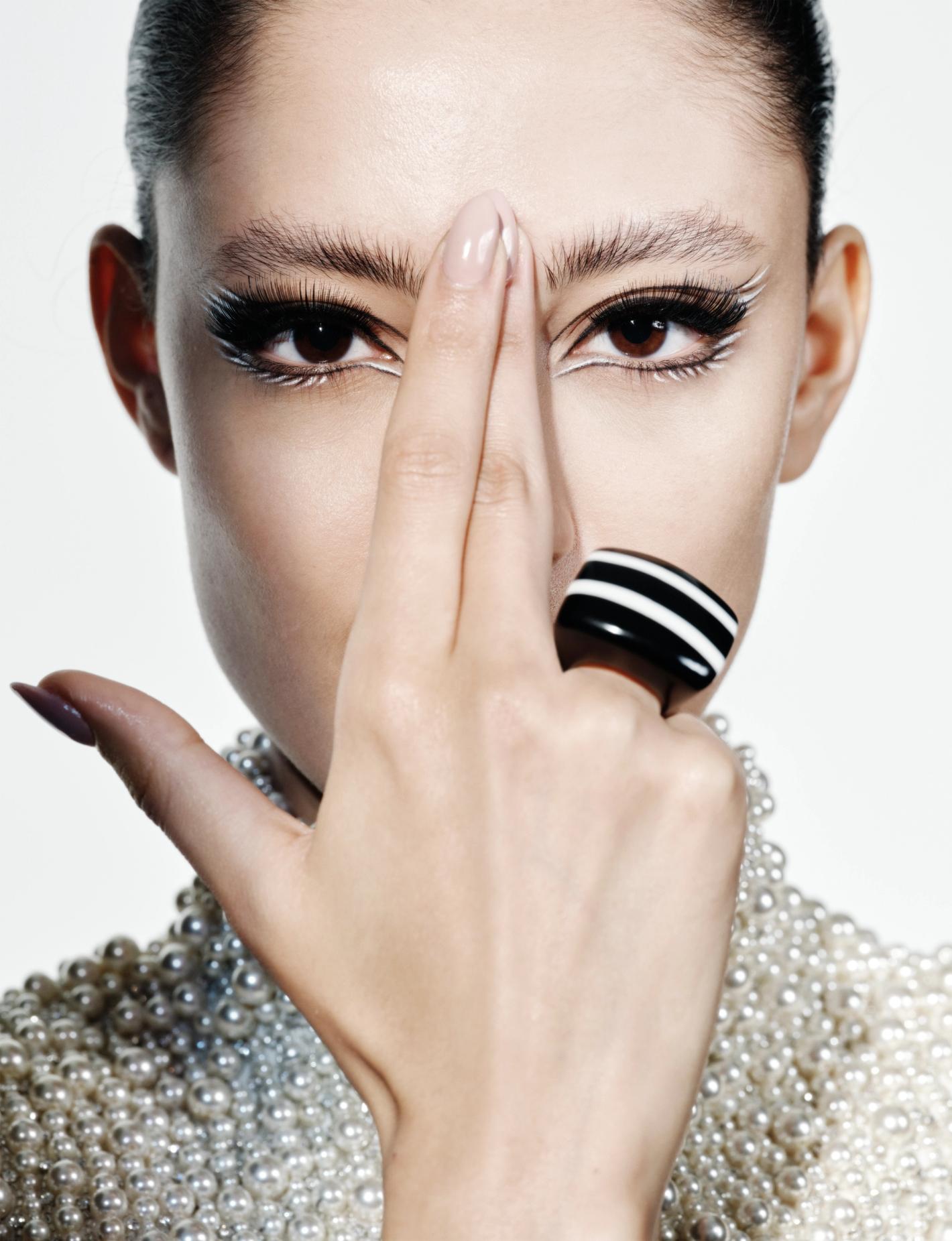 Close-up of a woman with elaborate white eyeliner and a pearl-beaded collar, posing with a black and white striped ring.