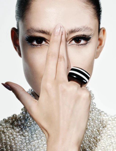 Close-up of a woman with elaborate white eyeliner and a pearl-beaded collar, posing with a black and white striped ring.
