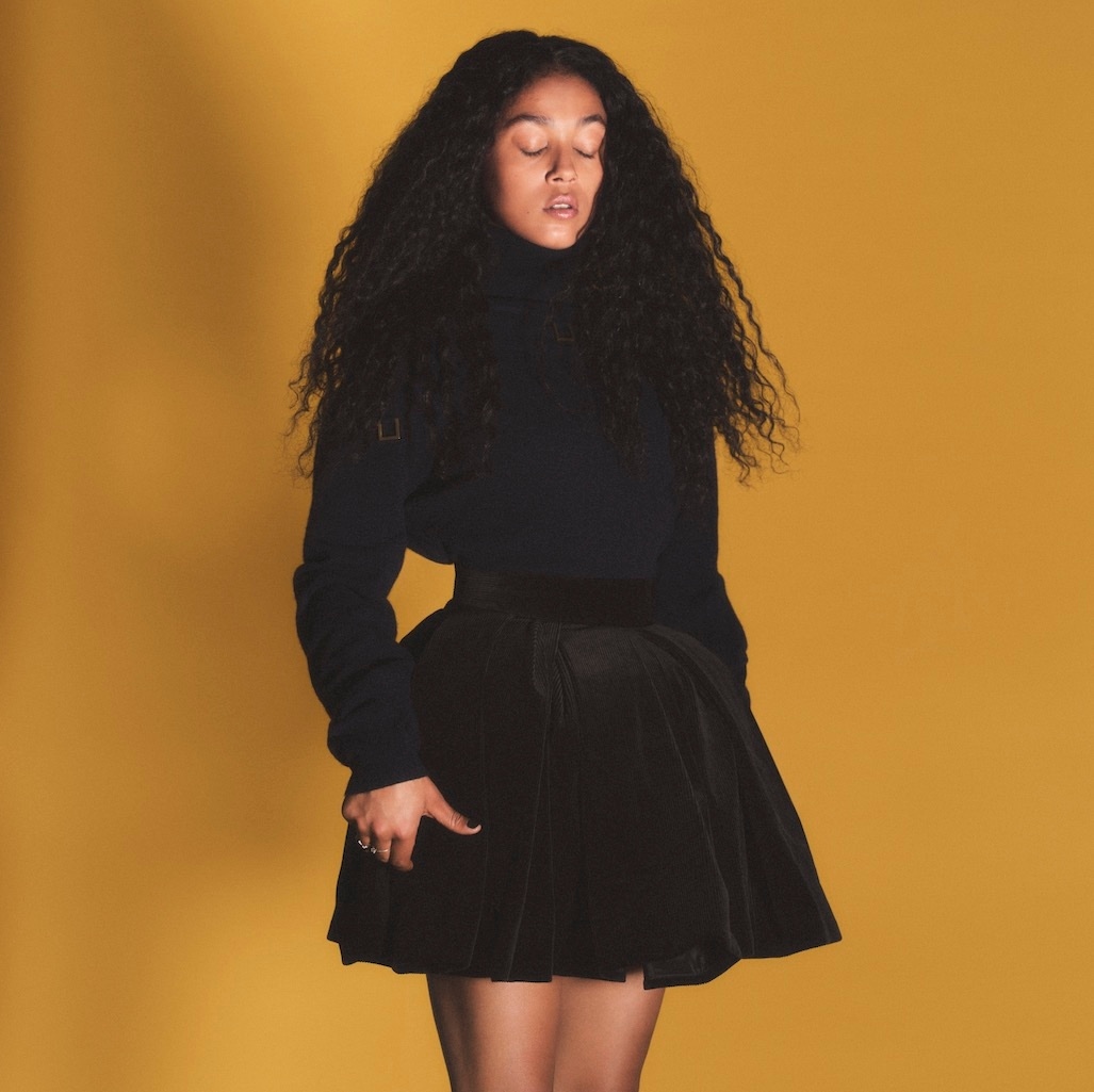 Elegant young woman with voluminous curly hair poses in a studio against a golden background, wearing a black turtleneck and skirt.