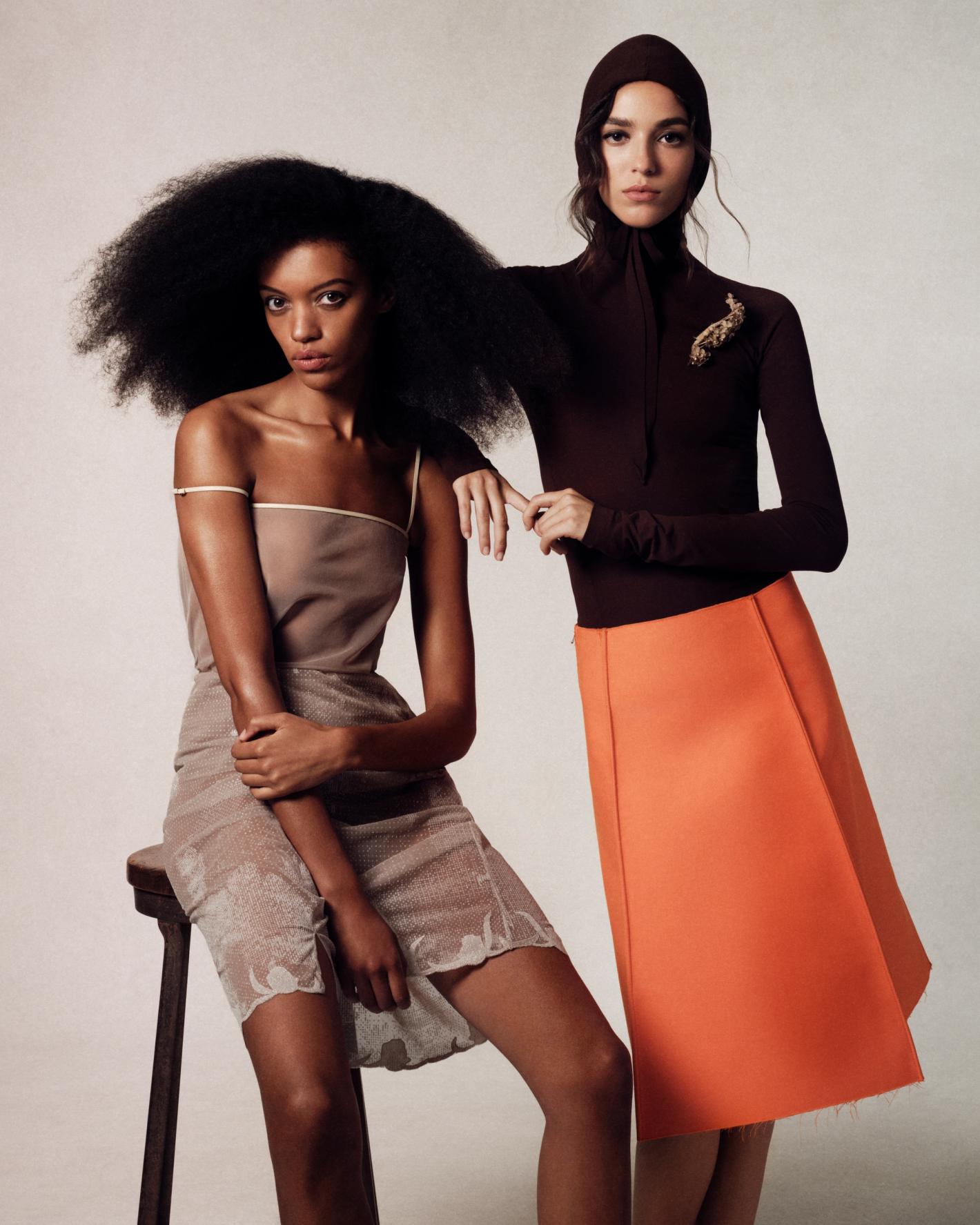 Two women pose in a studio wearing dresses. One woman sits on a stool wearing a brown dress. The other wears an orange skirt.