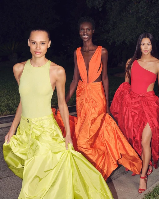 Three female models in vibrant gowns, walking on a path with greenery behind them, showcasing runway fashion with striking color contrasts.