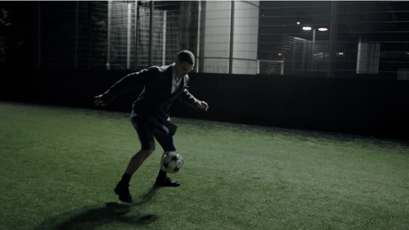Adult male in business suit balancing soccer ball on foot on outdoor astroturf field at night under stadium lights, action shot.