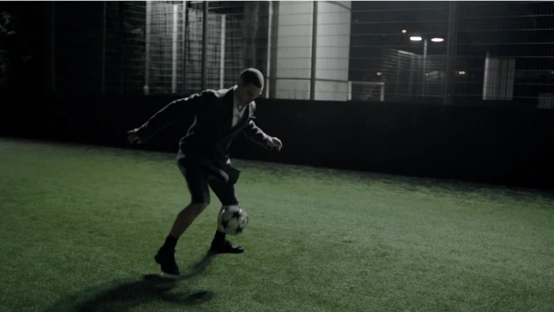 Adult male in business suit balancing soccer ball on foot on outdoor astroturf field at night under stadium lights, action shot.
