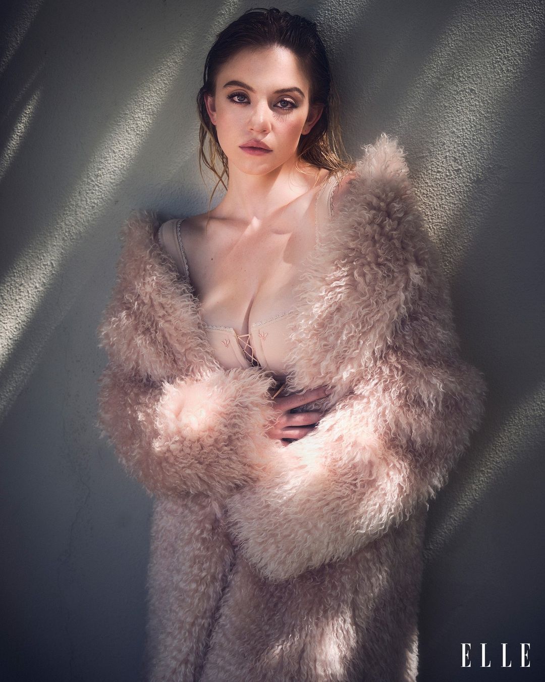 Sydney Sweeney poses in a pink fur coat against a textured wall with light and shadow, showcasing a fashion-forward editorial shot.