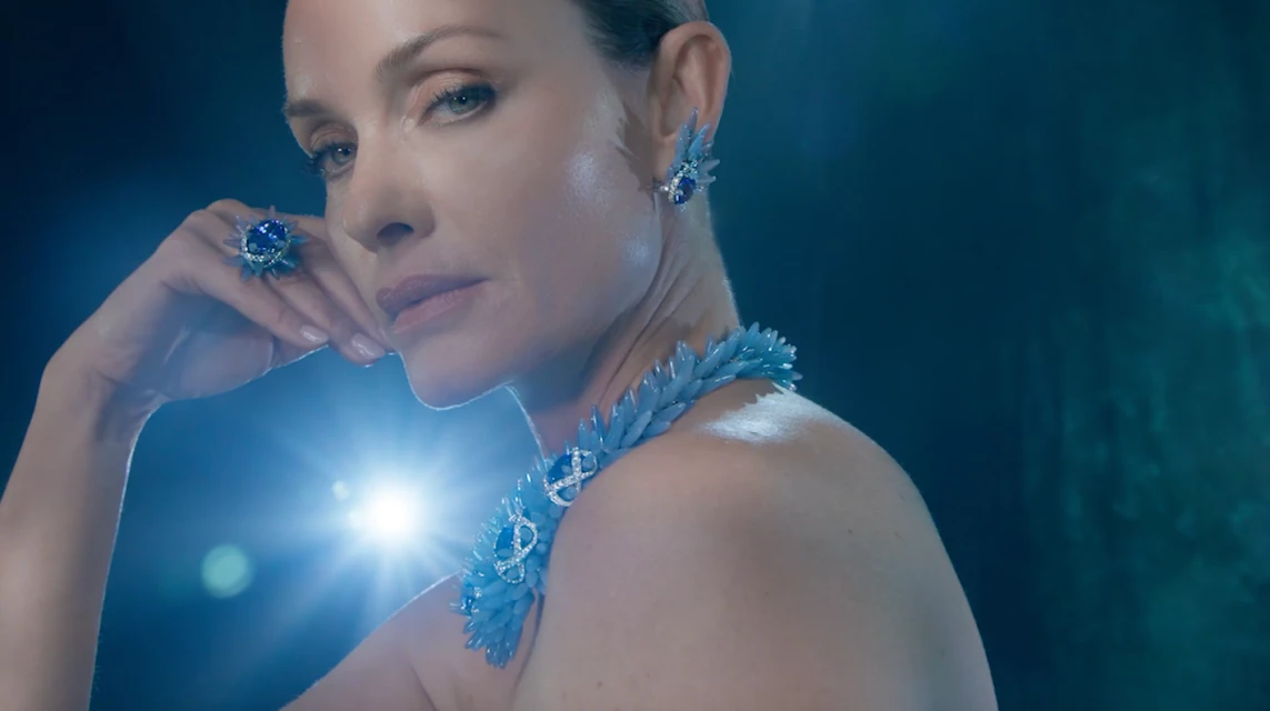 Woman poses wearing blue floral jewelry: necklace, earrings, and ring against a dark blue backdrop with bright studio lighting.