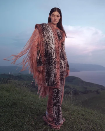 Model wearing a textured fringed knit outfit standing on a grassy hill overlooking a vast coastal landscape at dusk.