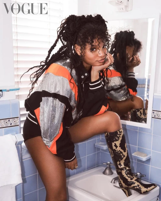 A woman with dreadlocks in a jacket and boots poses in a bathroom in front of a mirror in an indoor fashion editorial.