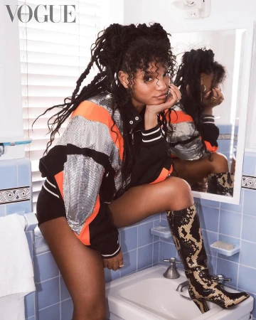 A woman with dreadlocks in a jacket and boots poses in a bathroom in front of a mirror in an indoor fashion editorial.