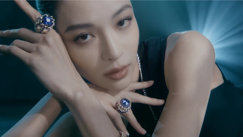 Close-up portrait of a woman wearing large blue gemstone rings and a necklace, posed against a glowing blue background.