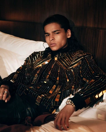 A man in his 20s wearing a heavily embellished gold and black patterned jacket seated on a hotel bed.
