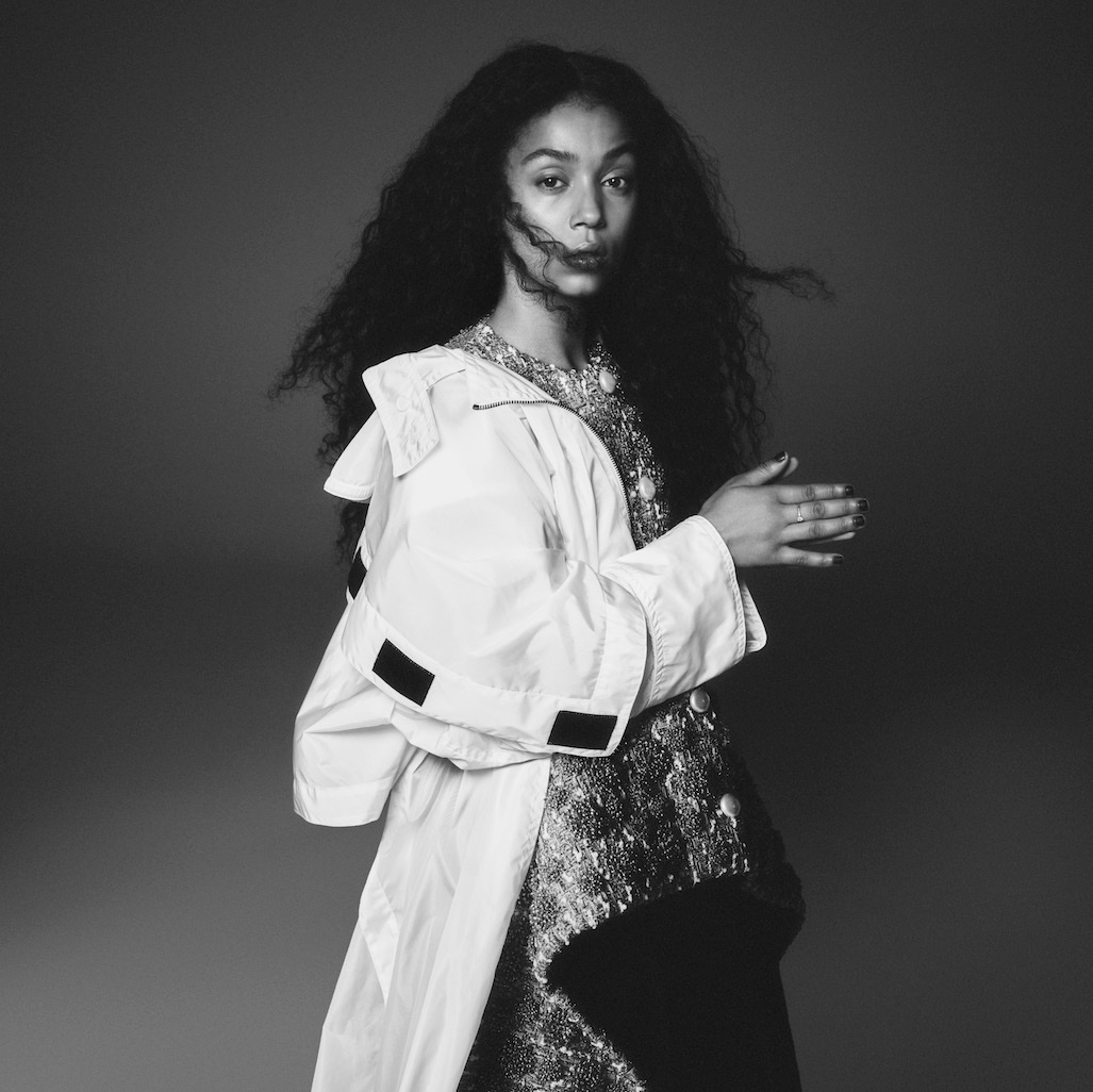 Black and white portrait of a stylish Black woman with curly hair wearing a white jacket and shimmering top in a studio setting.