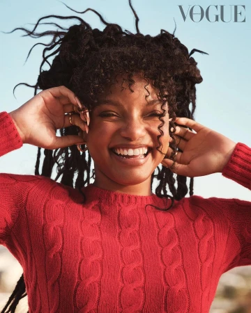 Smiling woman with dark curly hair wearing a red cable knit sweater against a clear sky