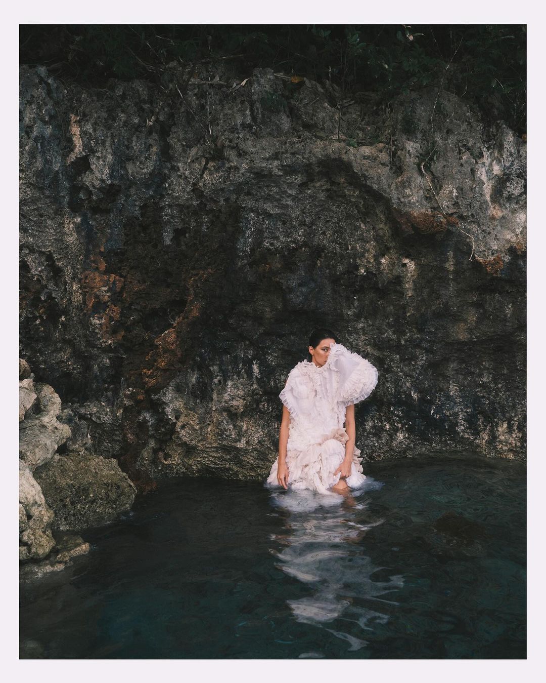 A woman in a white ruffled dress stands in water against a dark rock wall, creating a striking contrast in a natural setting.