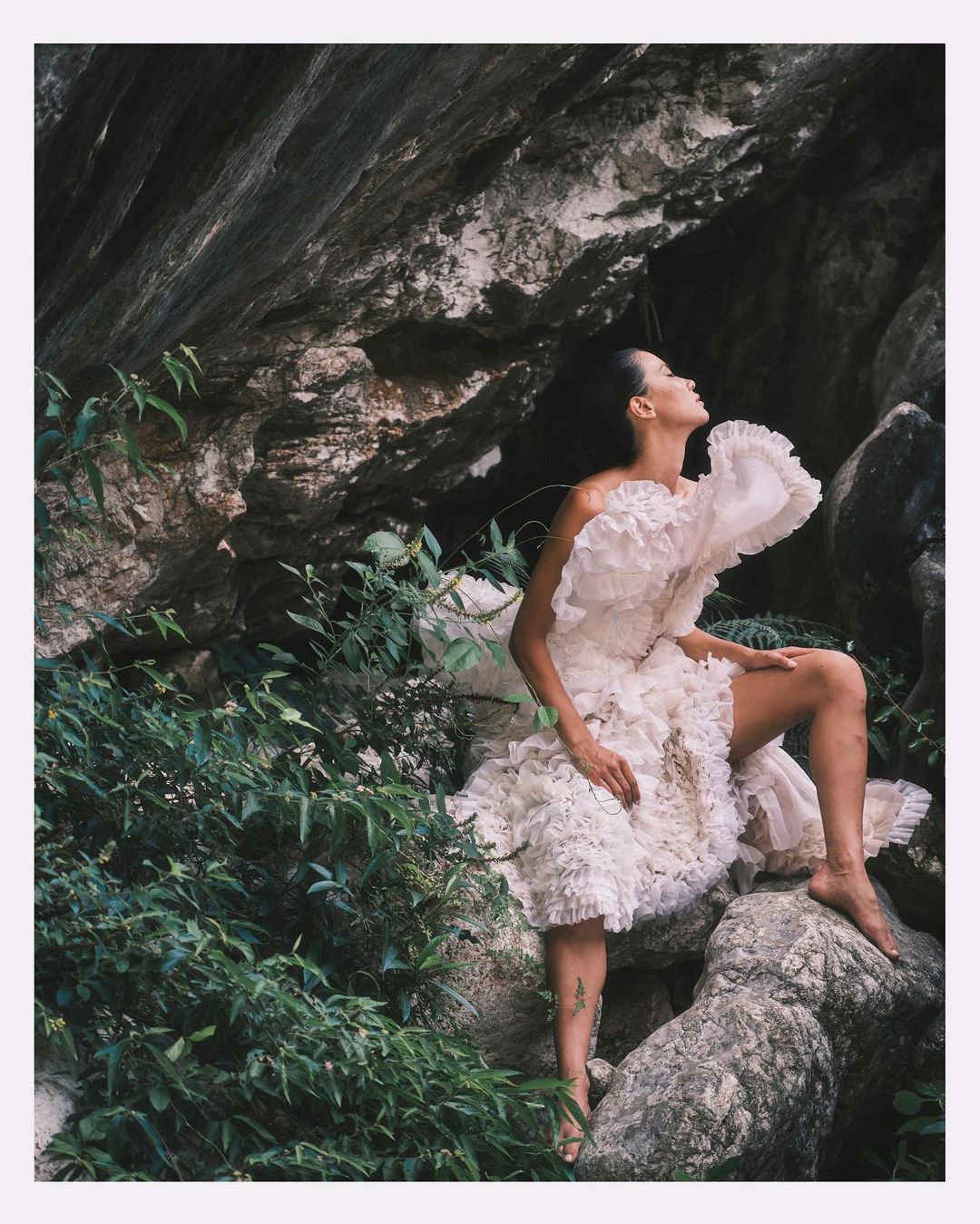Adult woman in a ruffled dress posing on rocks surrounded by lush greenery, illuminated by soft, natural light in an outdoor setting.