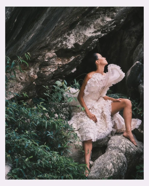 Adult woman in a ruffled dress posing on rocks surrounded by lush greenery, illuminated by soft, natural light in an outdoor setting.