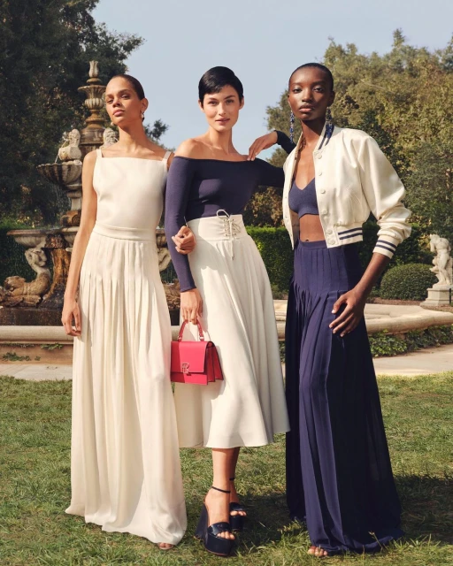 Three women model elegant spring fashions outdoors in an eye-level shot. They pose on grass near a water fountain, wearing dresses and heels.
