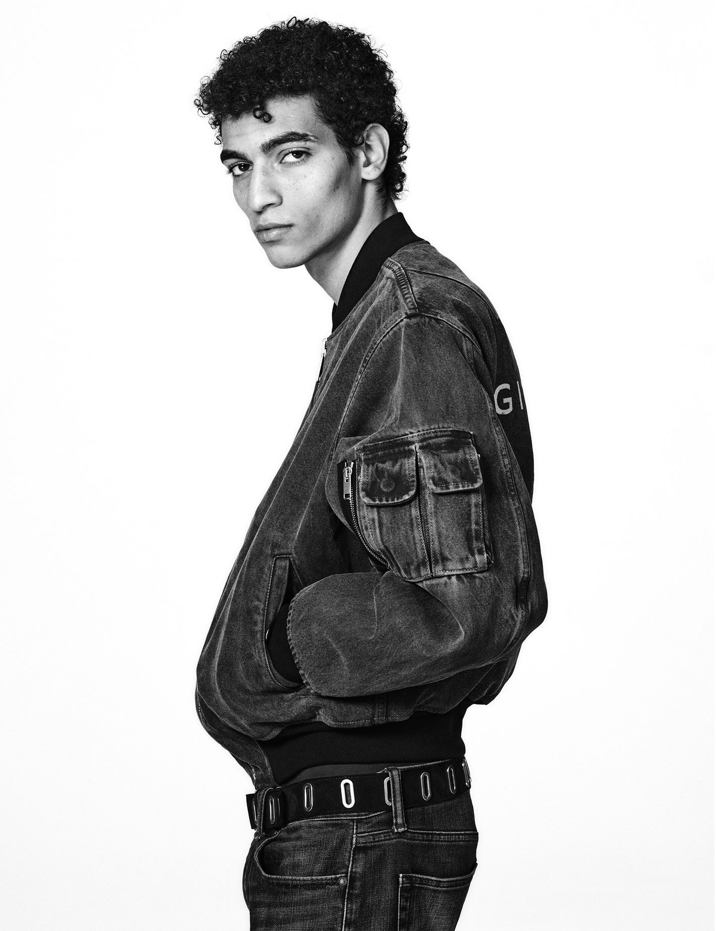 Adult model wearing a black coat, posing in a photo studio with a grey background and professional lighting. Fashion editorial portrait.