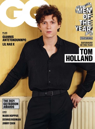 GQ magazine cover featuring Tom Holland in a pleated black shirt and matching pants, leaning against a light wood-paneled wall.