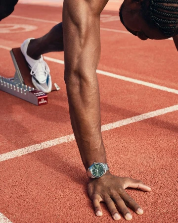 Close-up of an athlete in a crouched start position on a red running track, wearing a luxury wristwatch and white running shoes.