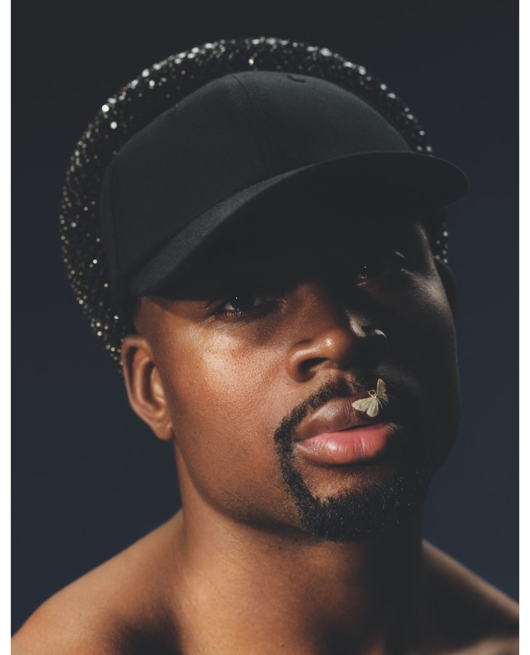 Close-up studio portrait of a stylish African-American adult male wearing a baseball cap, a sparkly hat, and with a butterfly on his lip.