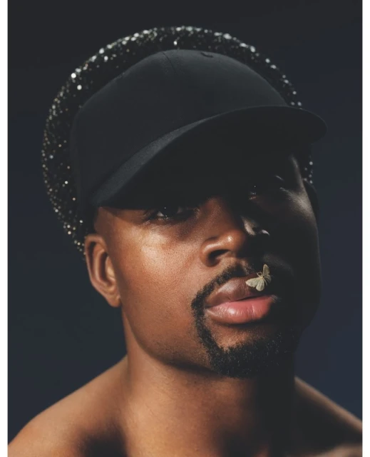Close-up studio portrait of a stylish African-American adult male wearing a baseball cap, a sparkly hat, and with a butterfly on his lip.