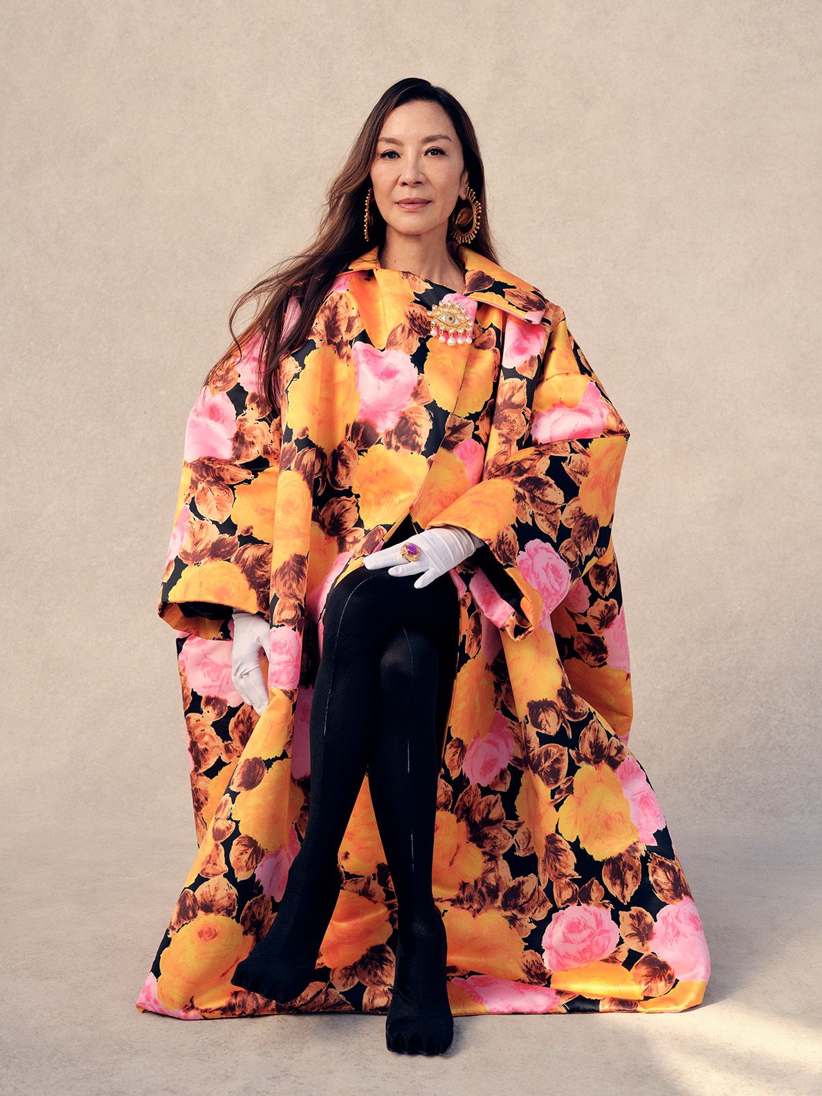 Michelle Yeoh poses in an oversized floral coat, black tights, and heels against a neutral background in a studio shot.