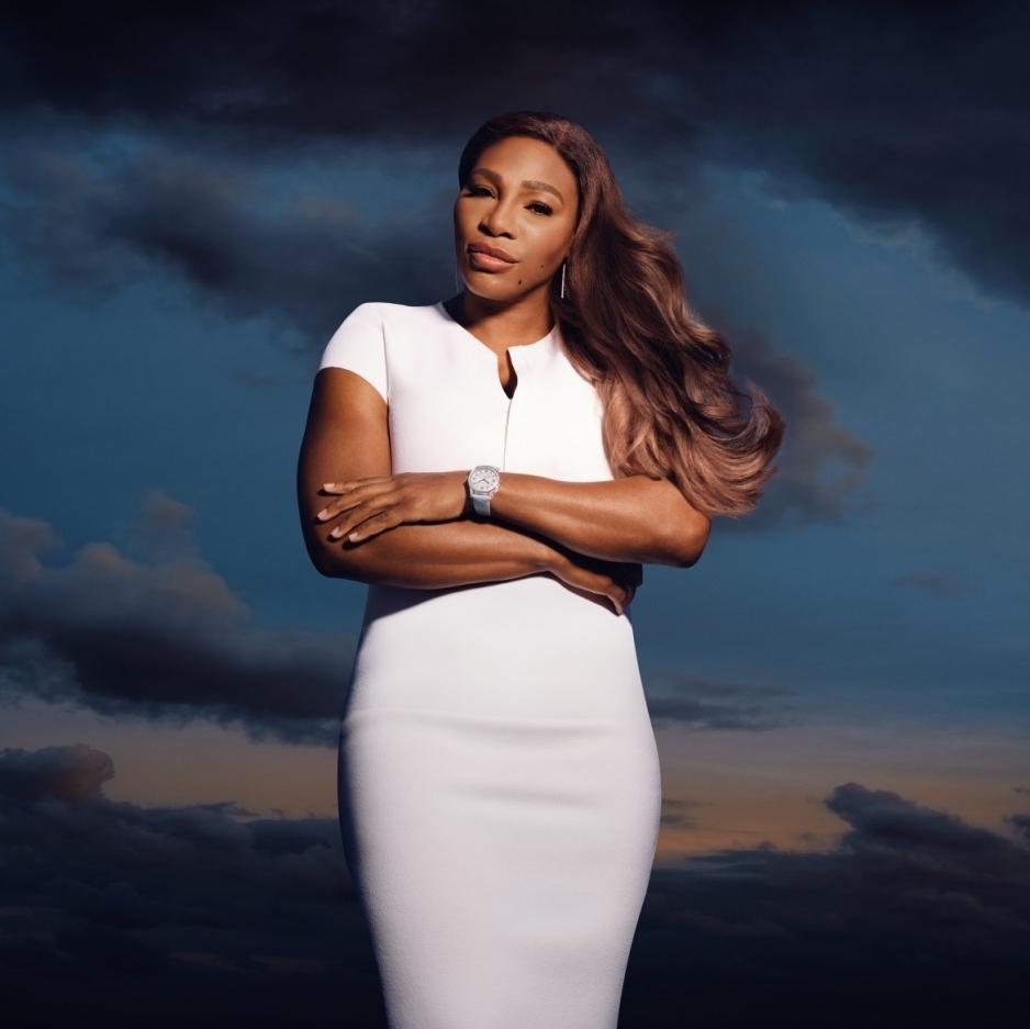 Portrait of an adult female in a white dress posing with arms crossed against a backdrop of clouds and sky at dusk.