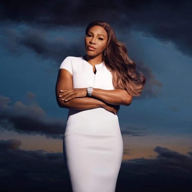 Portrait of an adult female in a white dress posing with arms crossed against a backdrop of clouds and sky at dusk.
