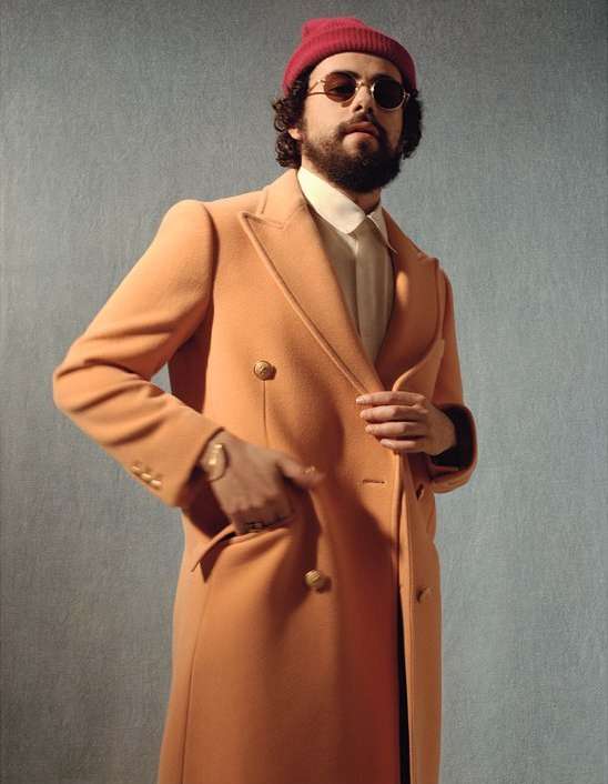Adult male wearing a red beanie, round sunglasses, a white shirt, and an orange double-breasted coat, posing in a studio environment.