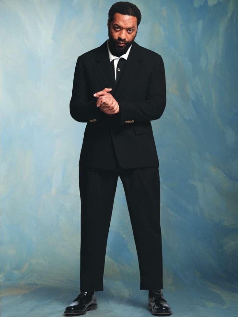 Full portrait of African American man in a black suit against a blue and white textured background, looking at the camera with hands together.