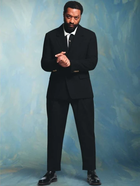 Full portrait of African American man in a black suit against a blue and white textured background, looking at the camera with hands together.