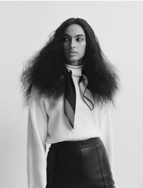 Portrait of a stylish adult woman with big, wavy dark hair, wearing a light sweater and a black skirt against a white backdrop.