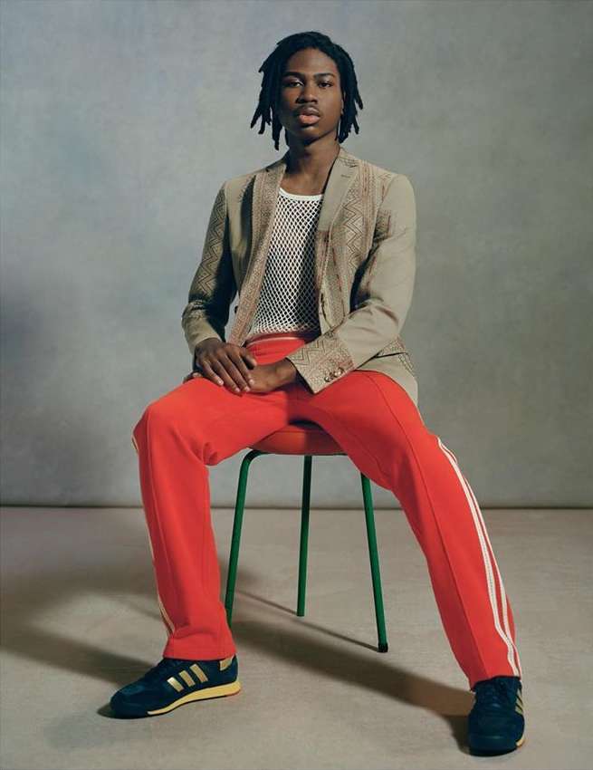 Young Black man sits on a green stool wearing a tan jacket, white mesh shirt, and red athletic pants in a studio portrait.