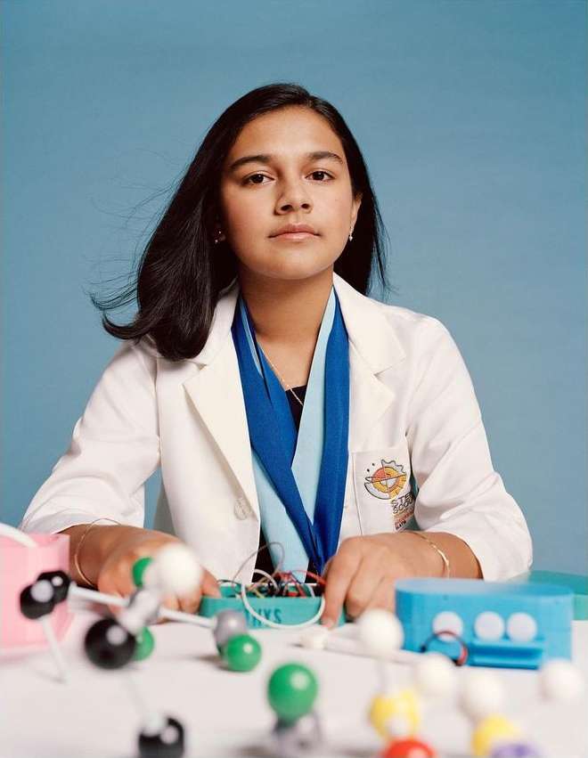 Portrait of a young woman in a lab coat seated at a table, surrounded by science equipment on a light blue background.