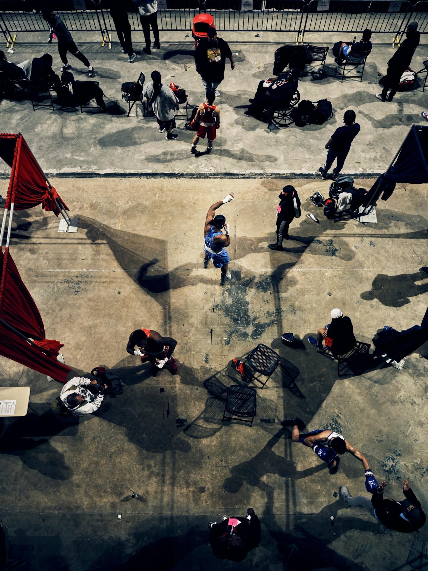 High-angle view captures a boxing match with two adult male boxers in red and blue shorts surrounded by spectators in an outdoor setting.