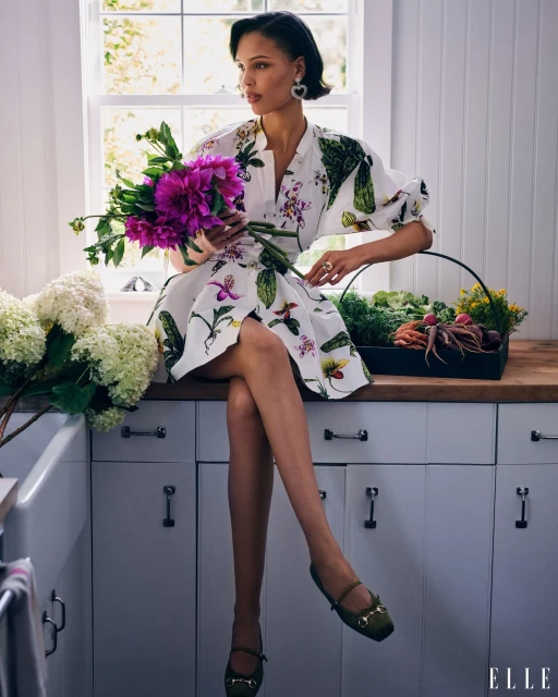 Adult female model in printed dress sits on white kitchen counter holding flower bouquet. Basket of root vegetables nearby. 