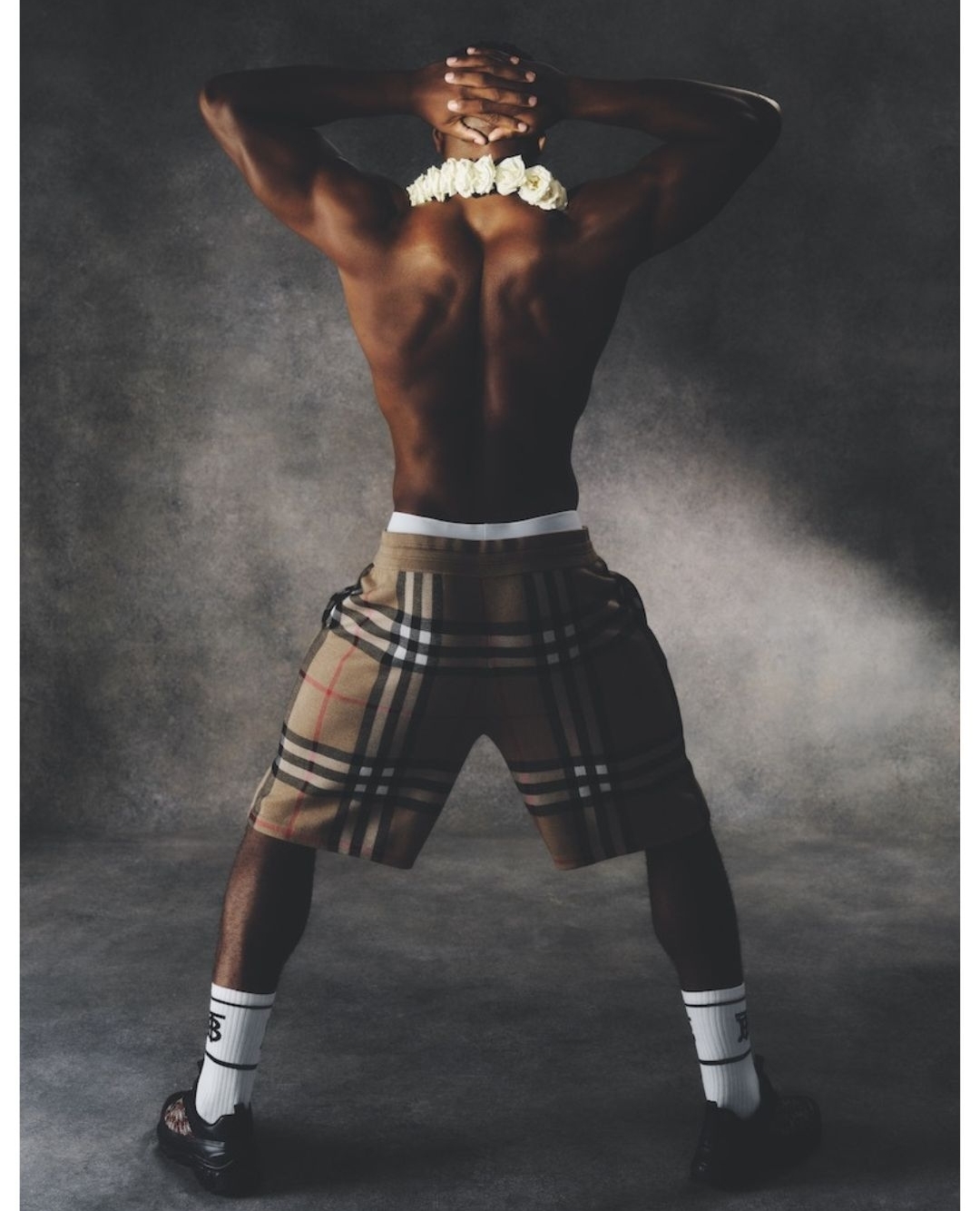 Man wearing Burberry plaid shorts and white socks poses with hands behind head. He has dark skin, muscular build, and white floral lei.